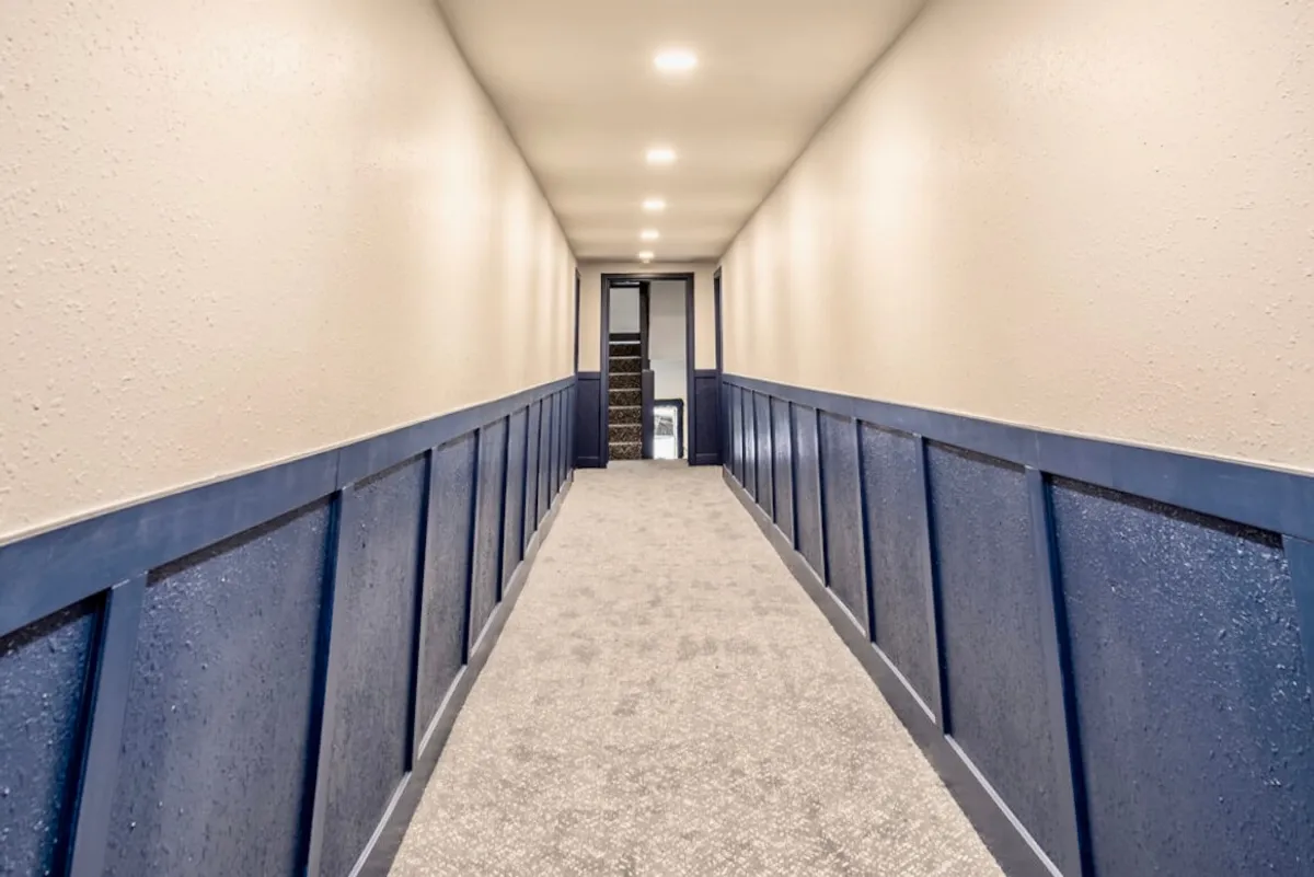 A welcoming hallway with blue wainscoting and a soft carpeted floor, leading toward a staircase at the end. The space feels calm and inviting, hinting at the anticipation of stepping outside or returning home after a day out in the city.