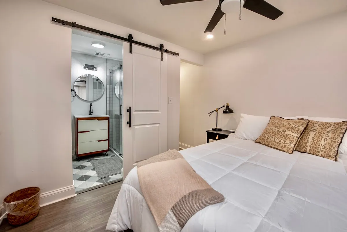 A tranquil bedroom scene featuring a plush bed adorned with decorative pillows, a warm table lamp on the nightstand, and a stylish sliding barn door that leads to the bathroom. Soft natural light filters in, creating a calming, inviting ambiance ideal for relaxation or reading.