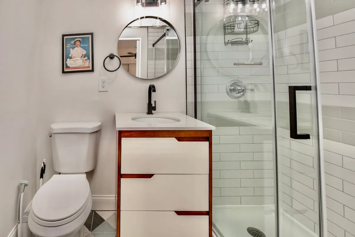 A bright, fresh bathroom featuring a round mirror above a sleek vanity with drawers, a walk-in shower with updated fixtures, and tasteful framed artwork. The space exudes a clean, modern feel, ideal for rejuvenation and personal care.