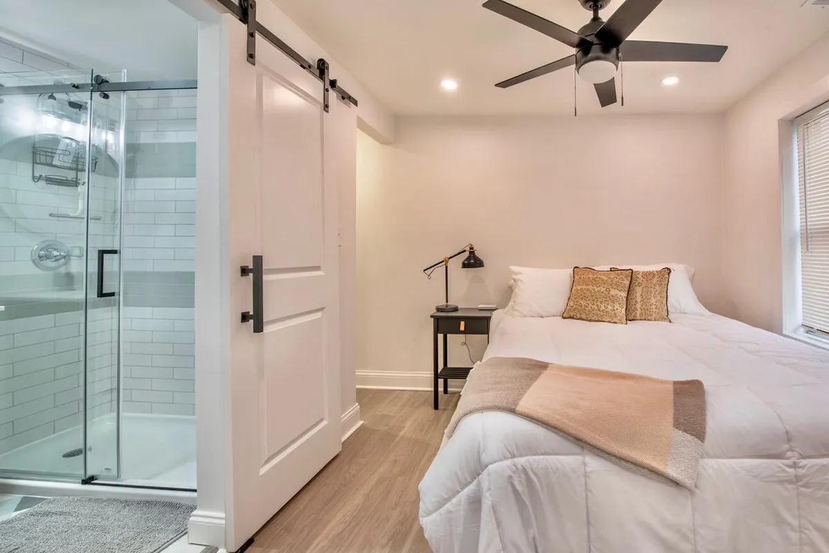 A serene angle that includes a comfortable bed with plush bedding, a nightstand with a softly glowing lamp, and a glimpse into the adjacent shower area. The room radiates warmth and peace, ideal for a restful night or an energizing morning routine.