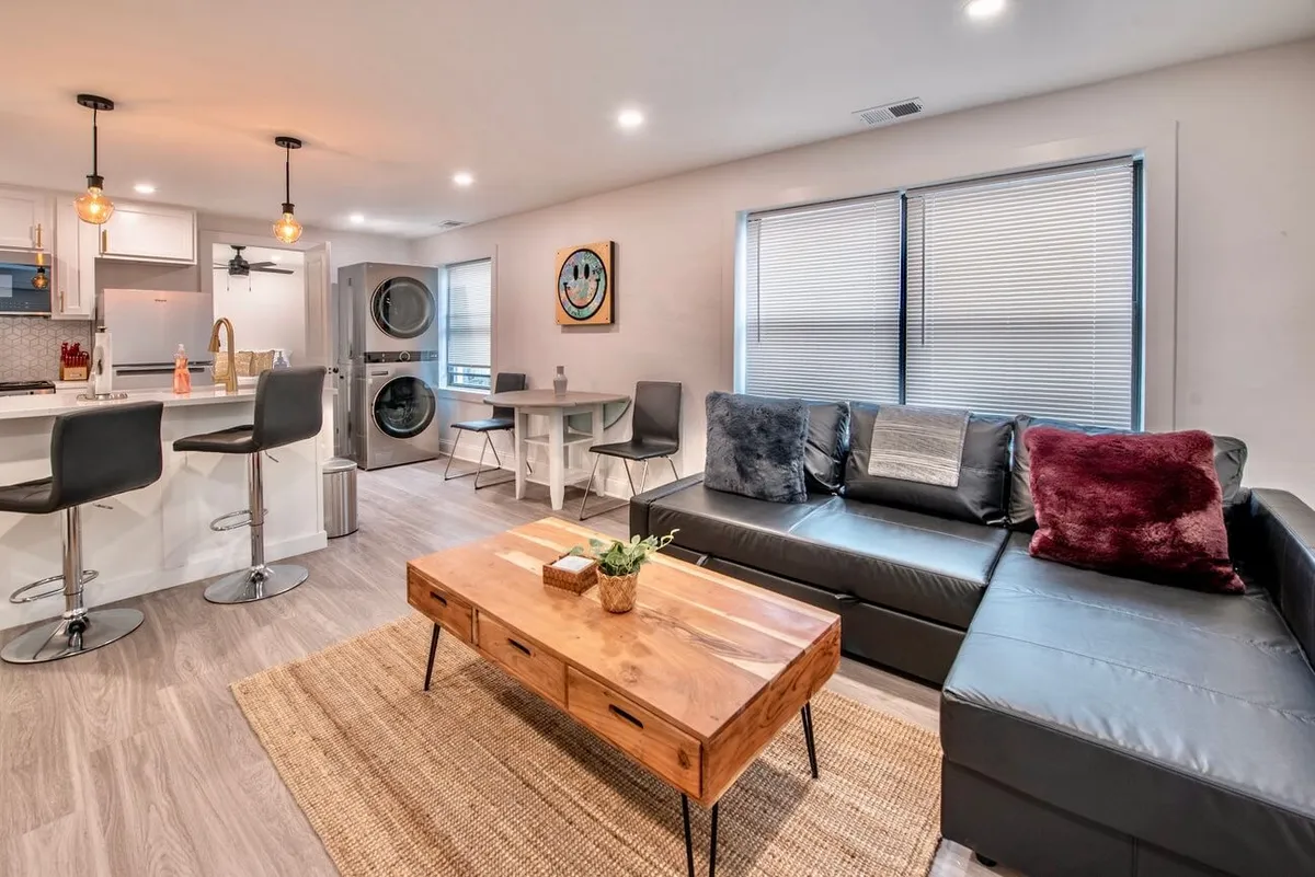 This image showcases a spacious open-plan living area featuring a black leather sofa, a modern wooden coffee table, and a kitchen island in the background. The room is accented by contemporary wall art, creating an inviting and stylish atmosphere. Ample light filters in, highlighting the clean lines and thoughtful layout ideal for both relaxation and socializing.